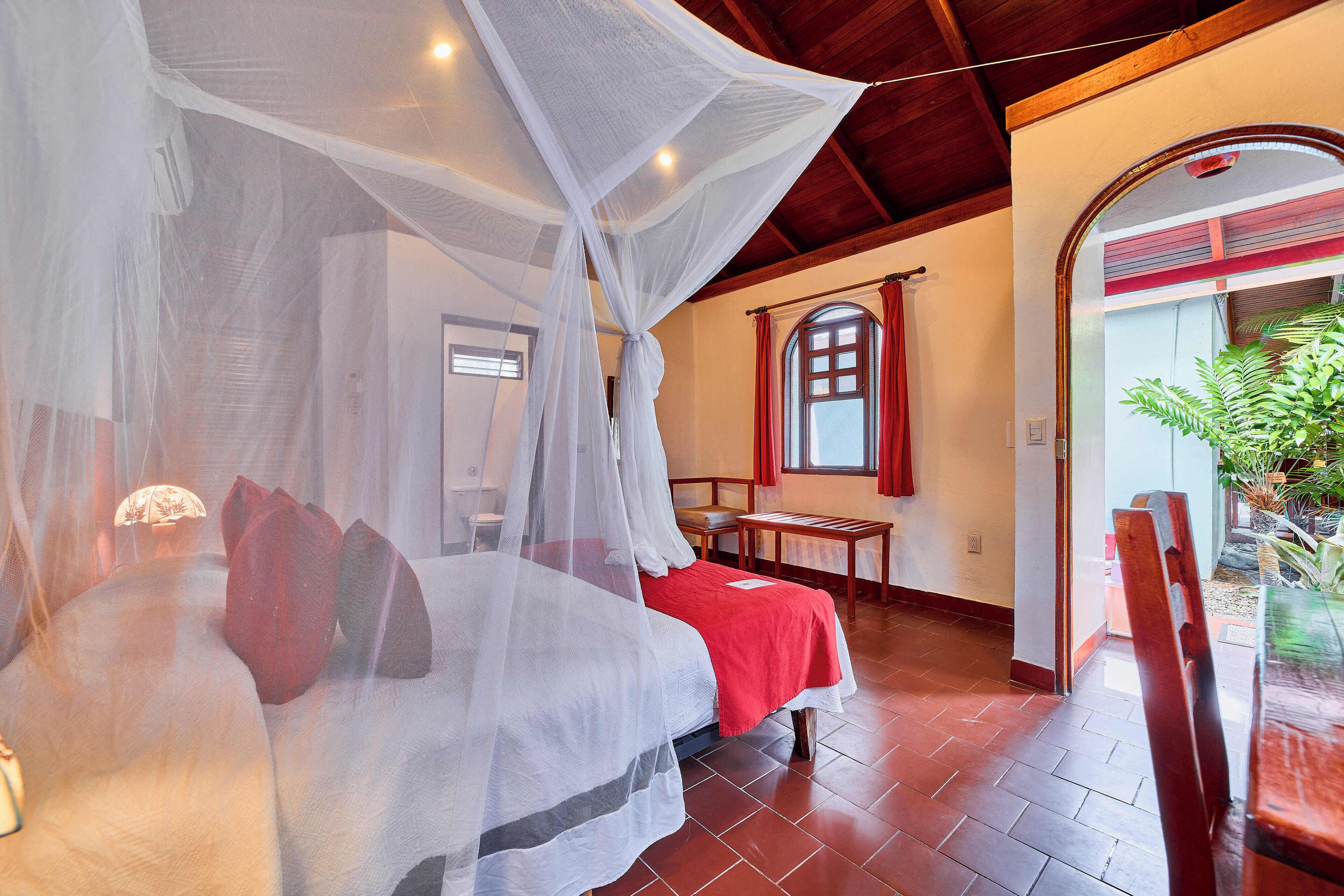 A bedroom with a bed under a mosquito net, a red bedspread, a wooden chair, and a window with red curtains.