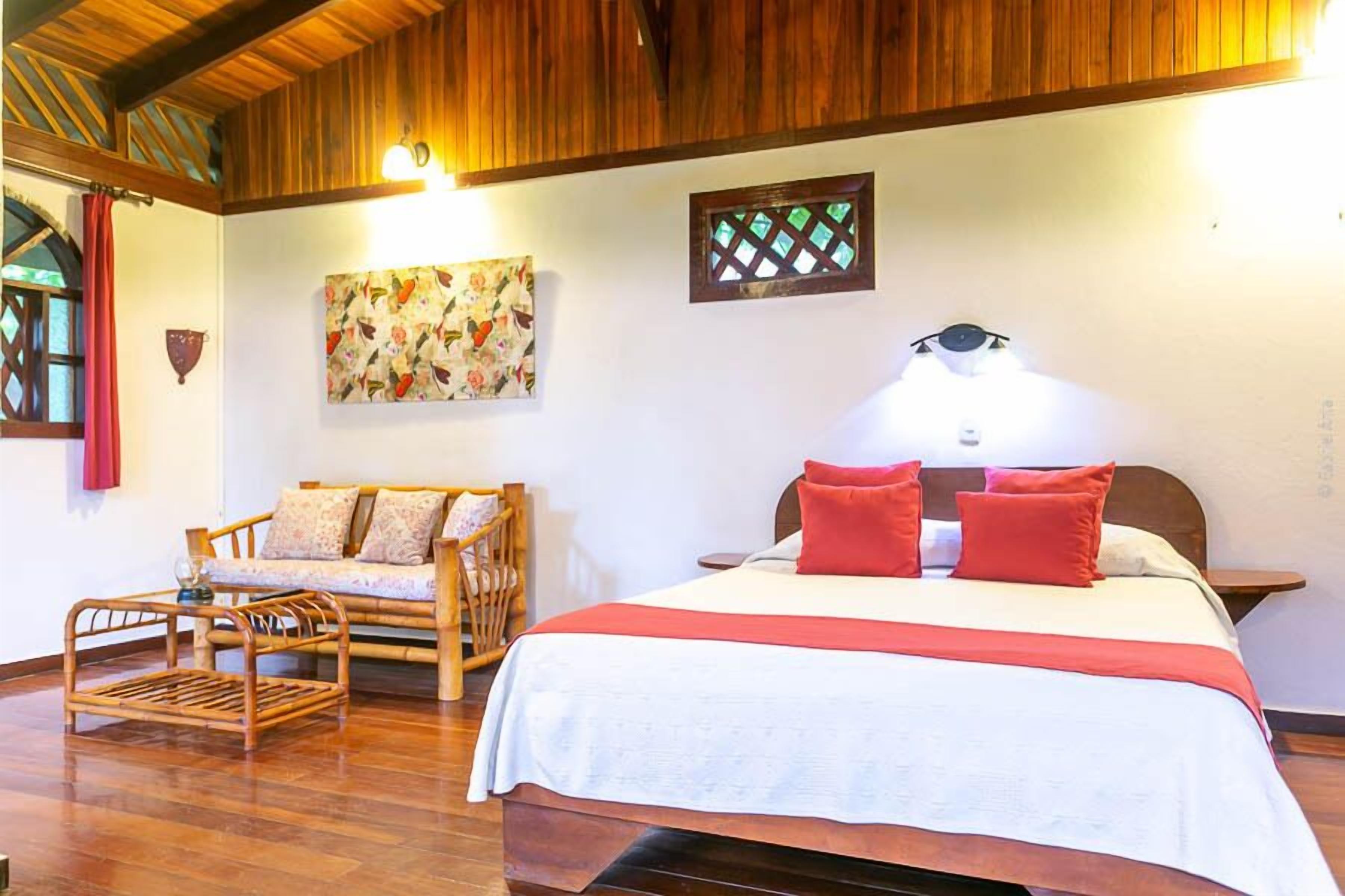 A bedroom with a wooden ceiling, a bed with red pillows, a wooden chair, a wall-mounted artwork, and a small window with a lattice design.