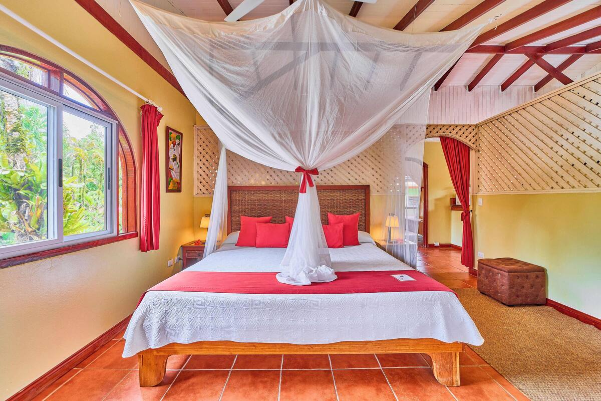 A spacious bedroom with a large bed, a canopy, and a view of greenery through a window.
