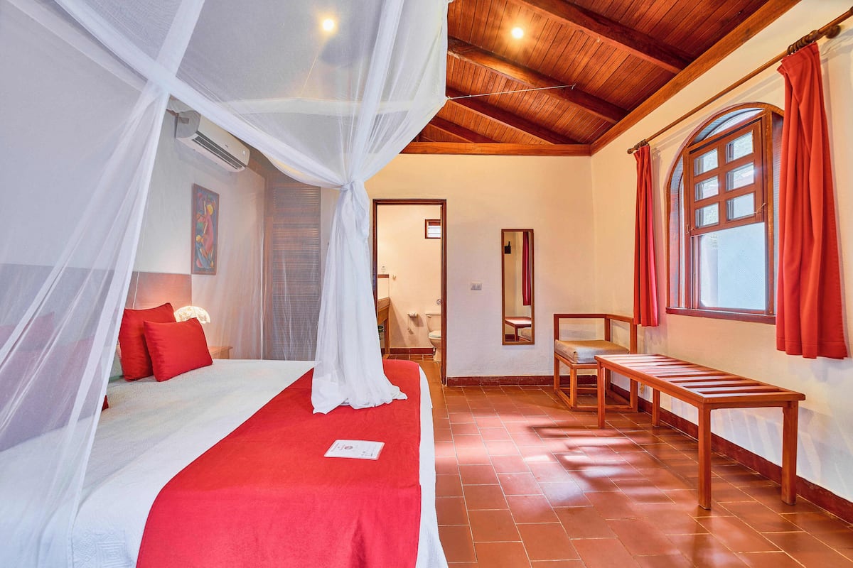 A bedroom with a bed, a wooden bench, a window with curtains, and a mirror on the wall.