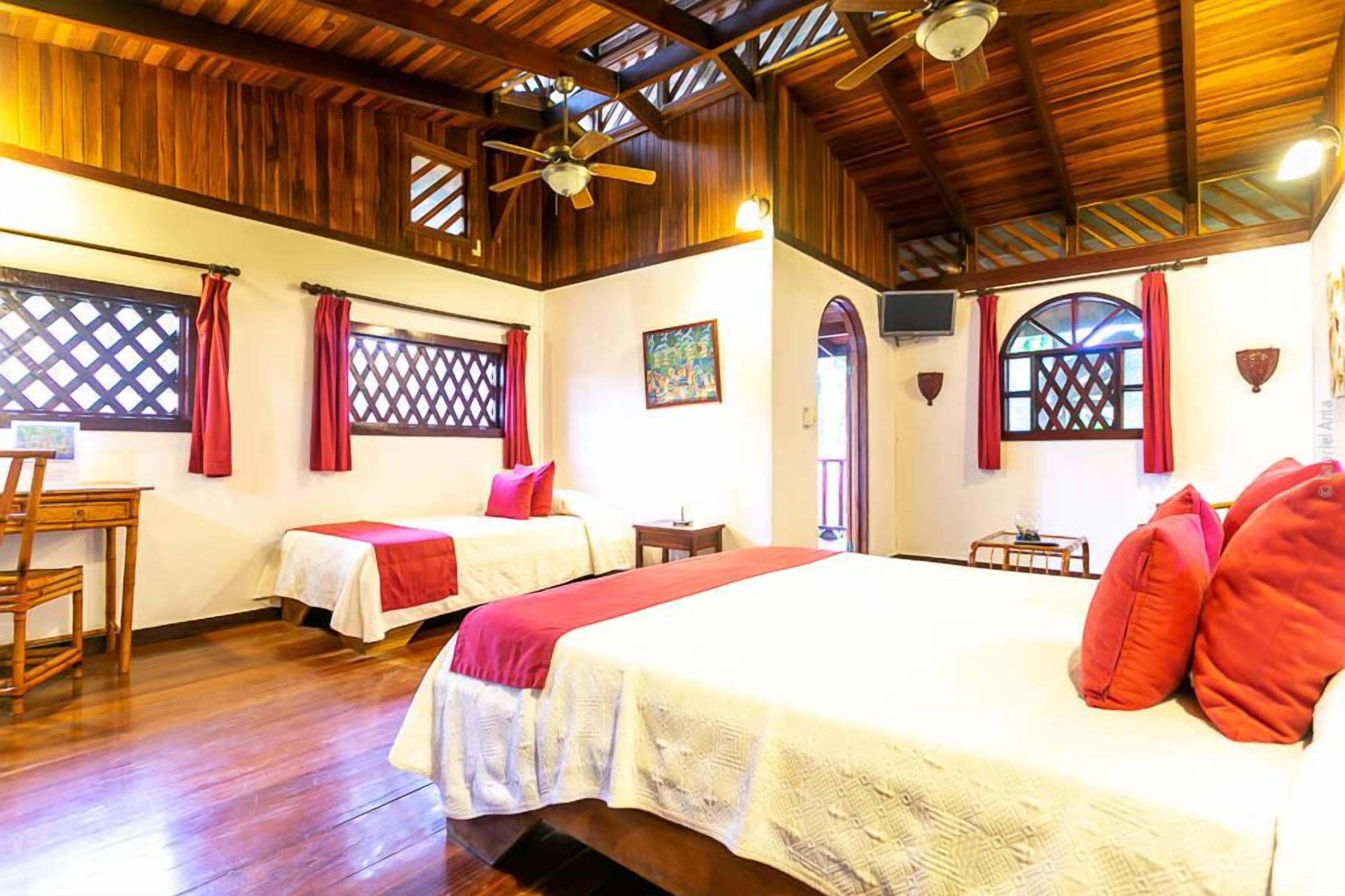 A room with two beds, wooden flooring, a ceiling fan, and a window with a lattice design.