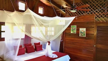 A wooden cabin bedroom with a bed under a mosquito net, red pillows, and two small windows.