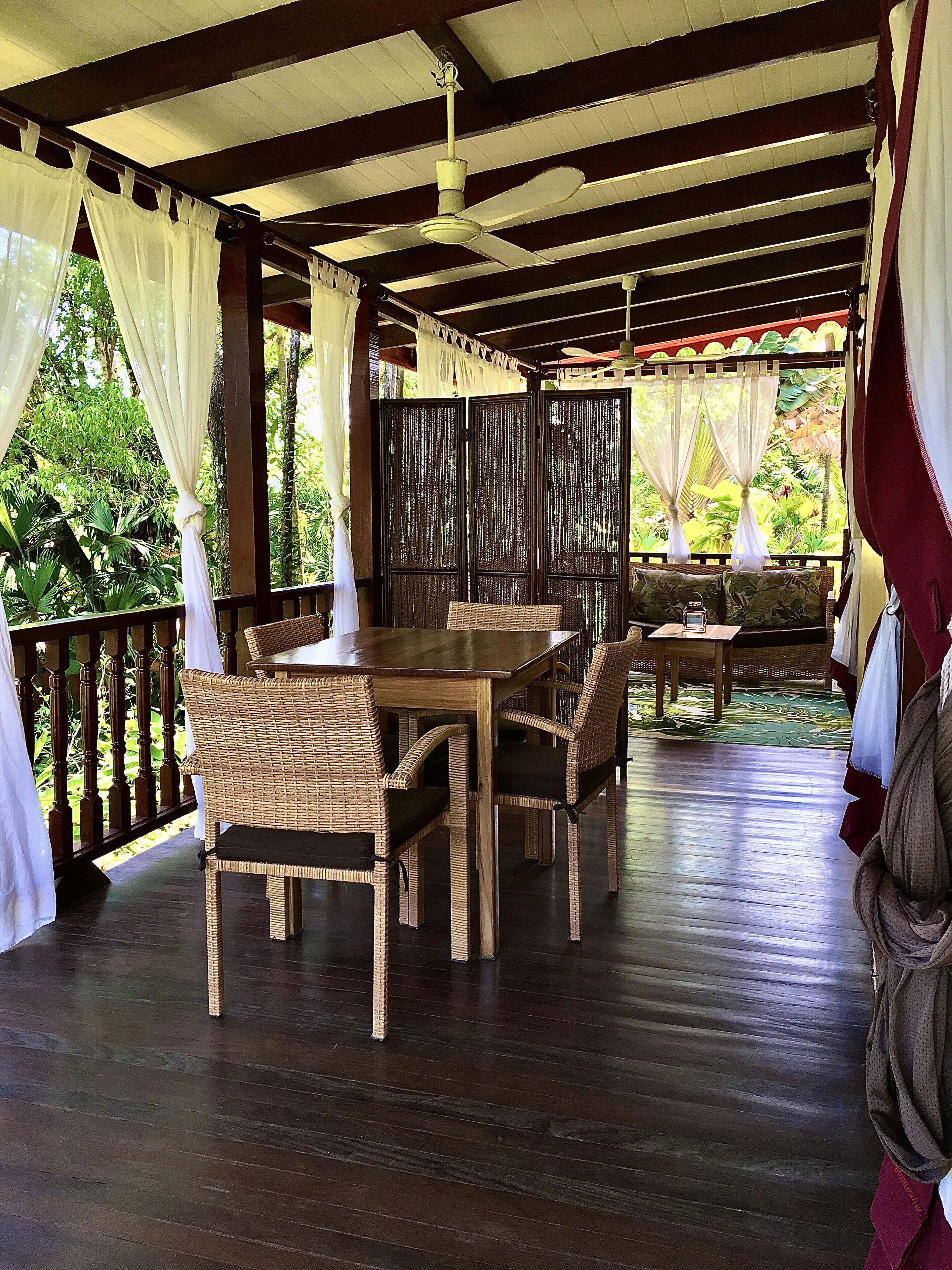 A covered outdoor dining area with a wooden table and wicker chairs, surrounded by a wooden railing and a view of lush greenery.