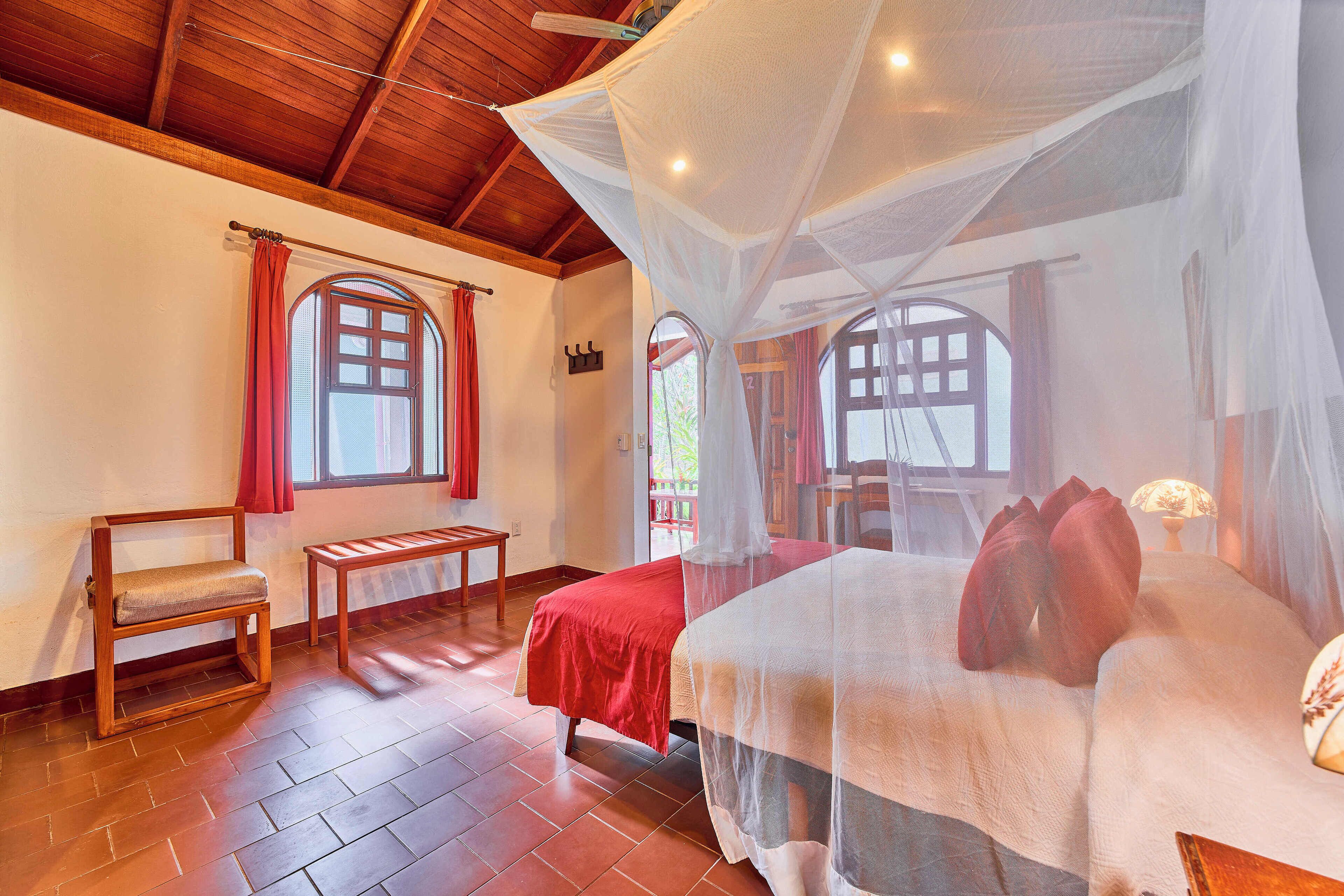 A room with a wooden ceiling, two beds with red and white bedding, a bench, and a window with red curtains.