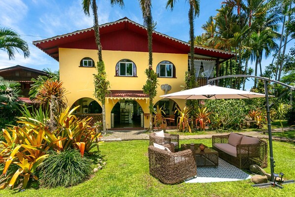 A yellow building with a red roof, surrounded by tropical plants and a patio area with wicker furniture. - Hotel El Encanto (Cahuita)