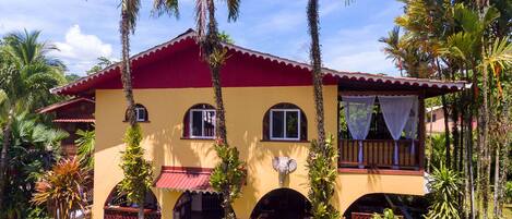 Un edificio de dos plantas con techo rojo y paredes amarillas, rodeado de palmeras y plantas tropicales.