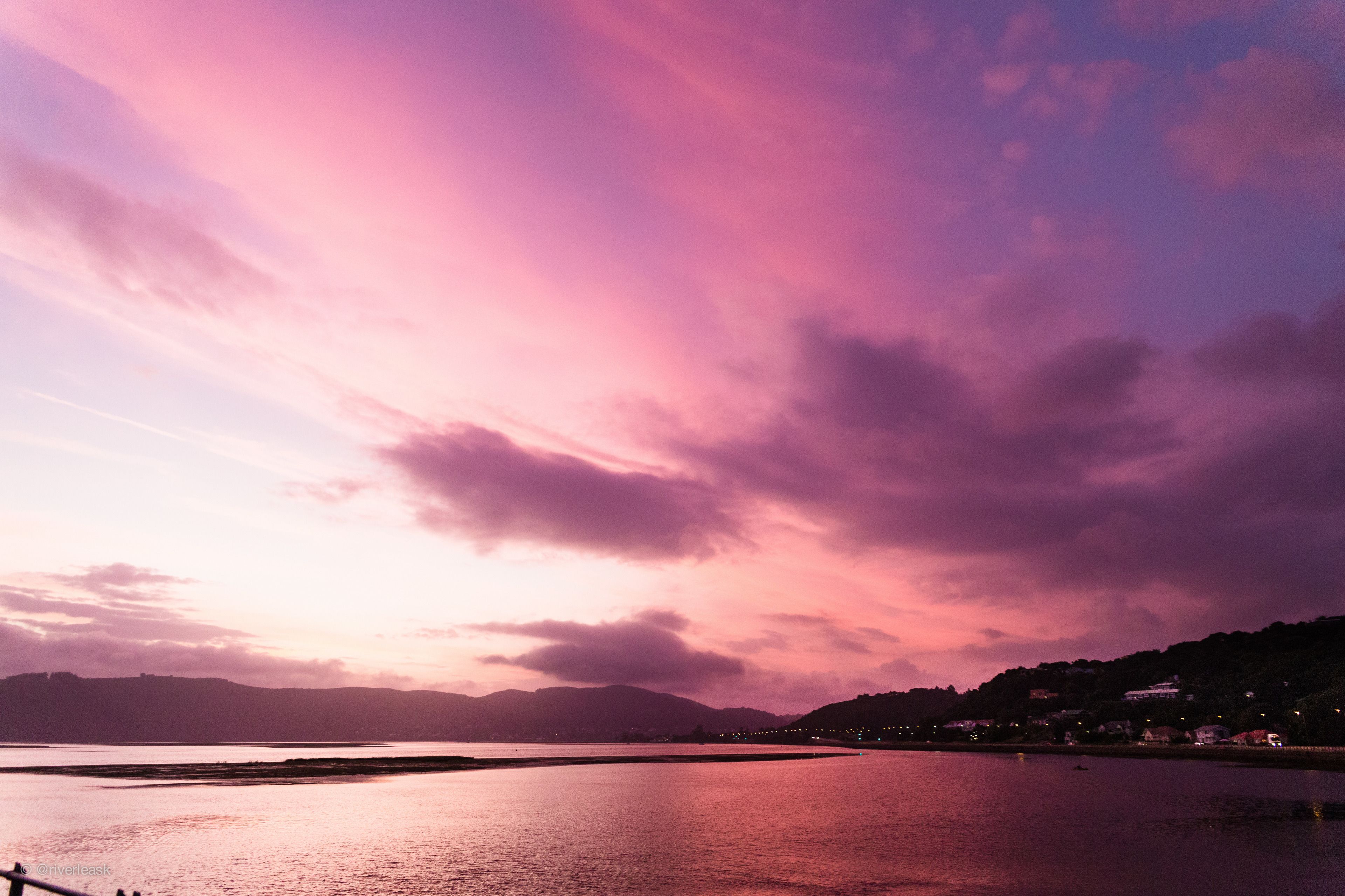 A sunset over a calm body of water with distant hills and a few buildings on the shore.