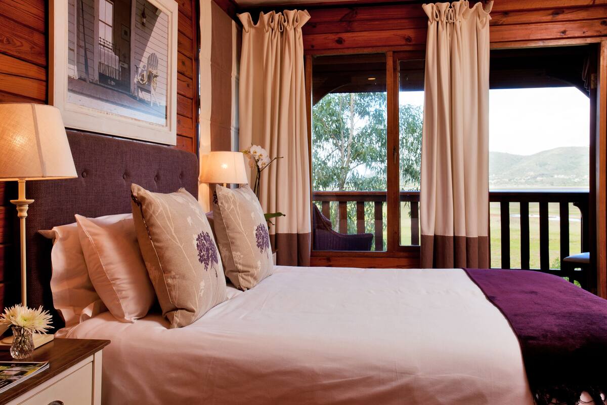 A bedroom with a bed, two bedside lamps, a window with curtains, and a view of greenery outside.