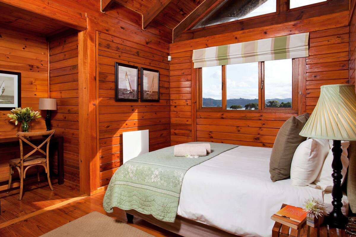 A wooden cabin bedroom with a bed, pillows, a bedside lamp, a chair, a desk, and a window with a view.