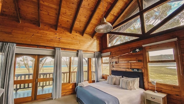 A wooden cabin bedroom with a bed, nightstand, and large windows offering views of a grassy field.