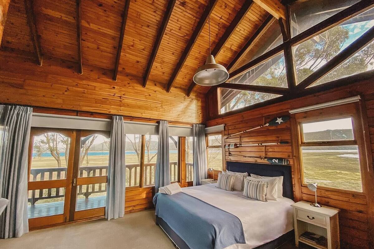 A wooden cabin bedroom with a bed, nightstand, and large windows offering views of a grassy field.