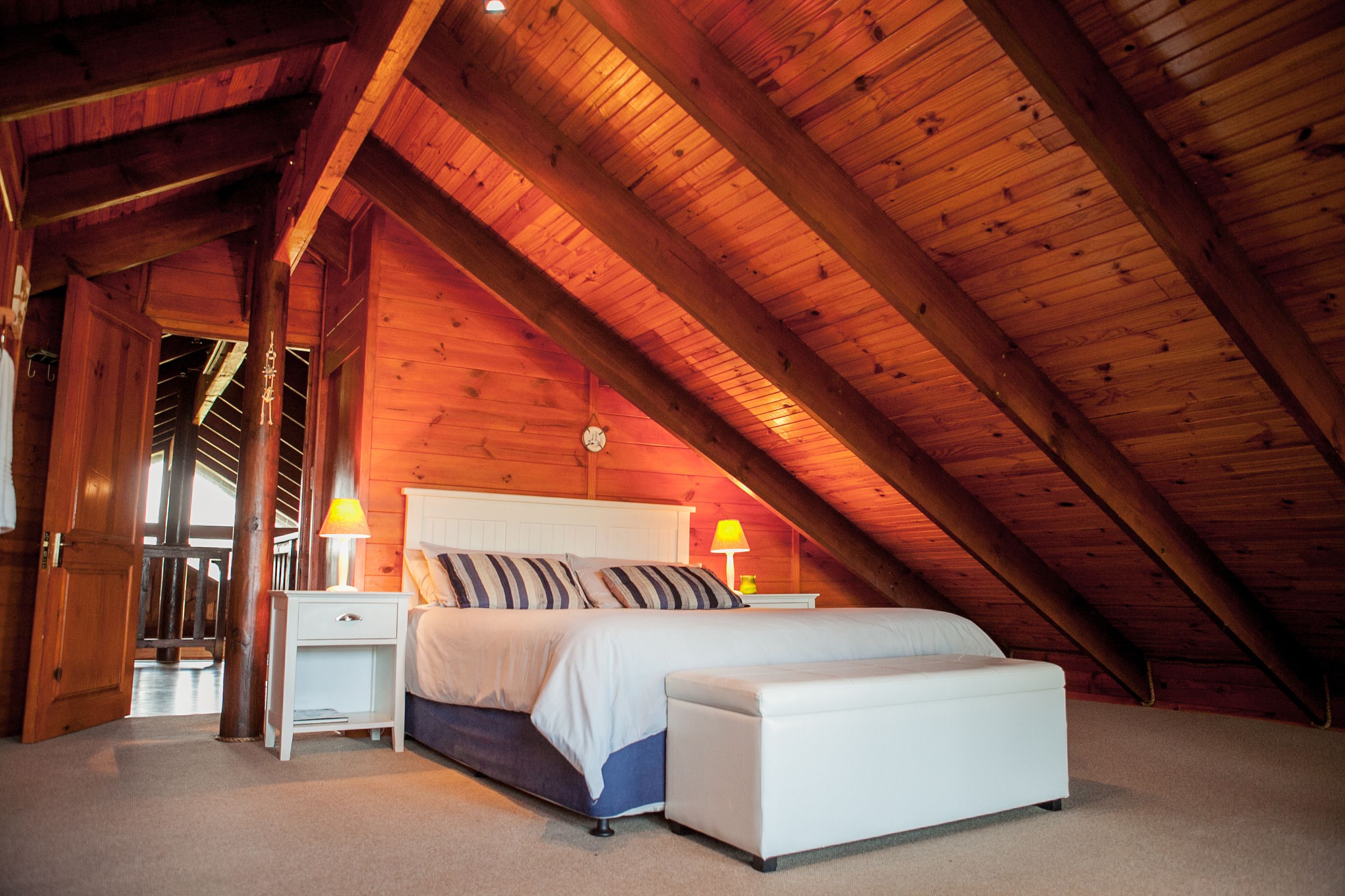 Ein Schlafzimmer mit schrägen Wänden, Holzbalken, einem Bett mit weißer Bettwäsche, zwei Nachttischen mit Lampen und einer Sitzbank am Fußende des Bettes.
