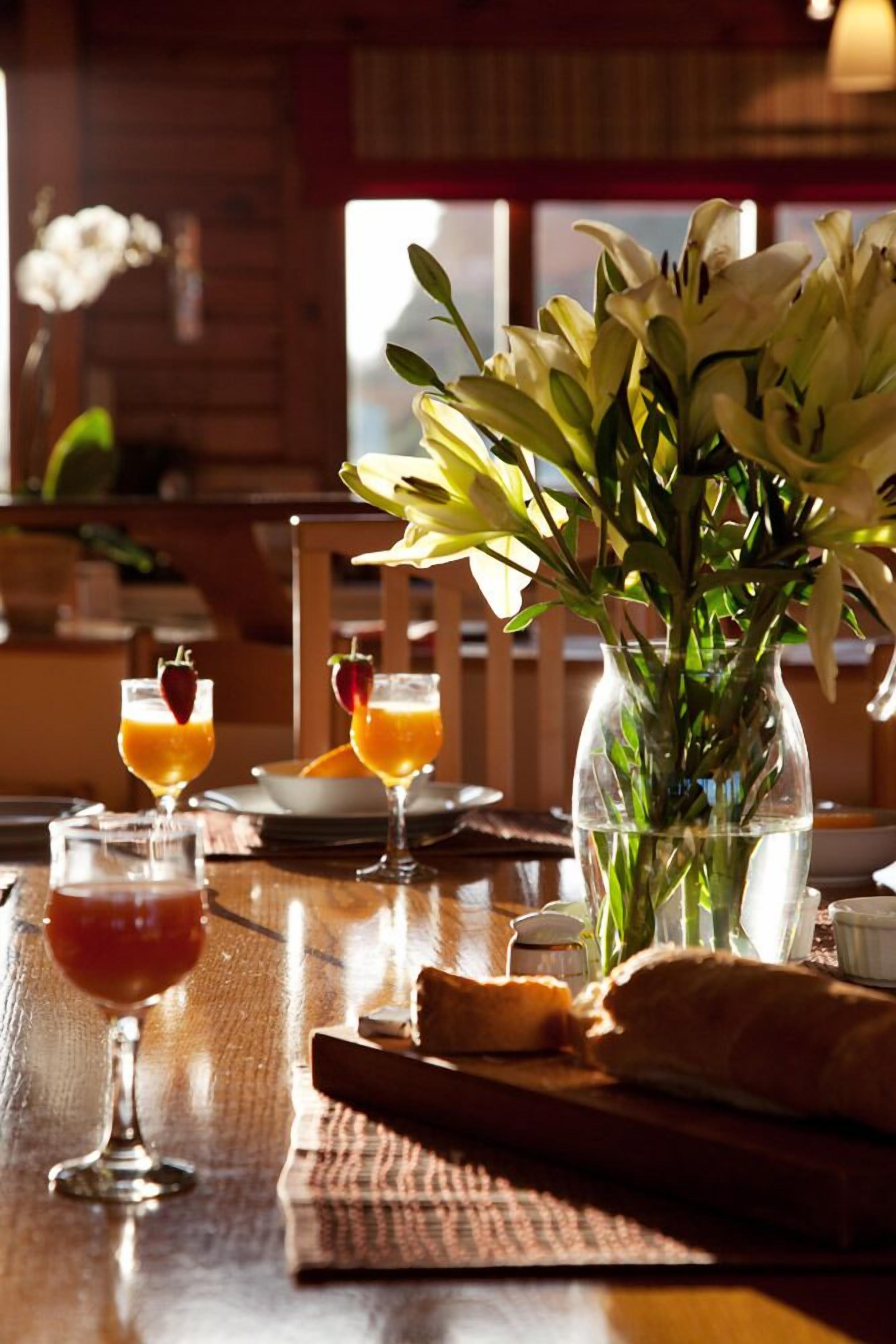 Una mesa de comedor de madera con un jarrón de lirios blancos, dos vasos de zumo de naranja con fresas y una hogaza de pan.