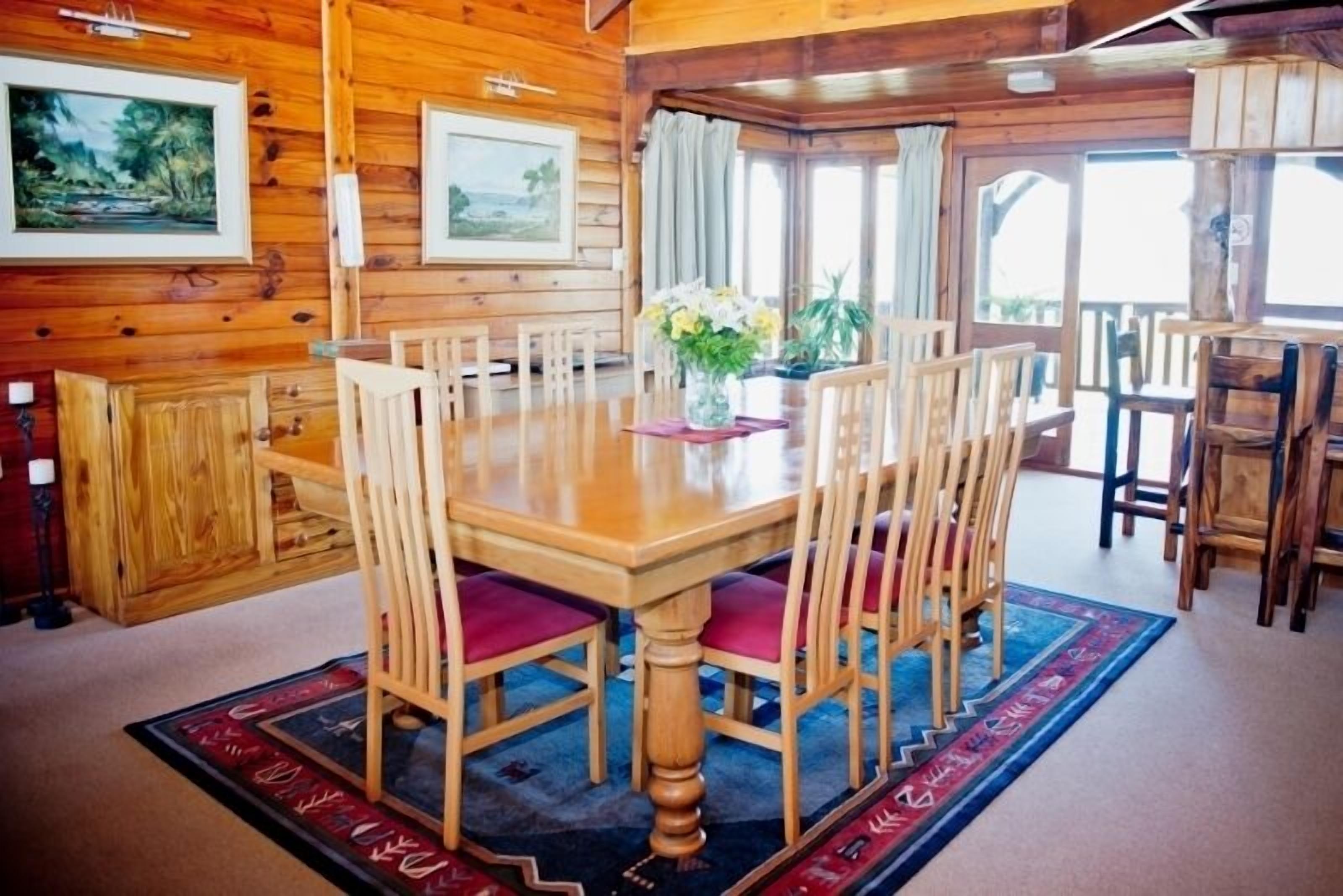 A wooden dining area with a large table and chairs, framed artwork on the walls, and a view through the windows.