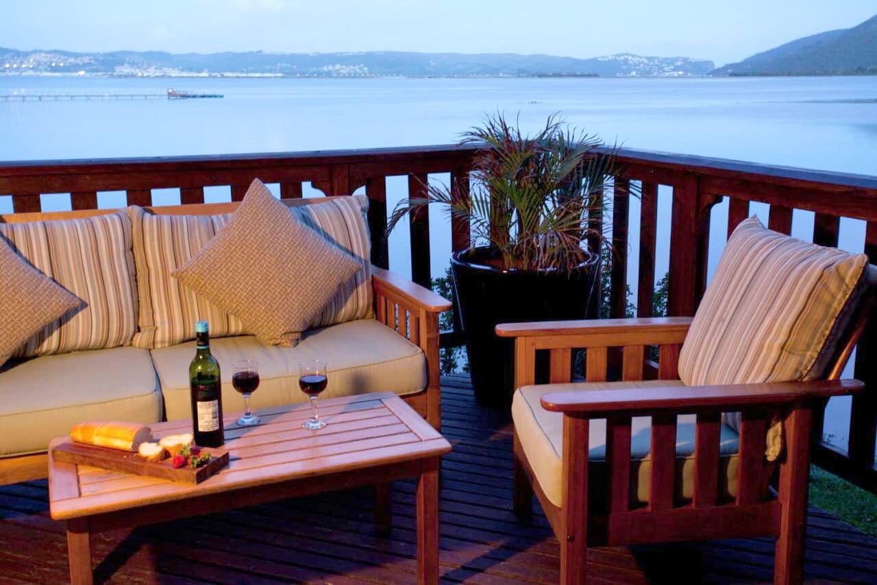 Una terraza de madera con un sofá y sillones, una mesa con una botella de vino y copas, y una planta en maceta.