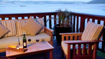 Una terraza de madera con un sofá y sillones, una mesa con una botella de vino y copas, y una planta en maceta.
