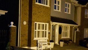 Une maison à deux étages avec un porche à l’avant, un banc et une voiture garée dans l’allée.