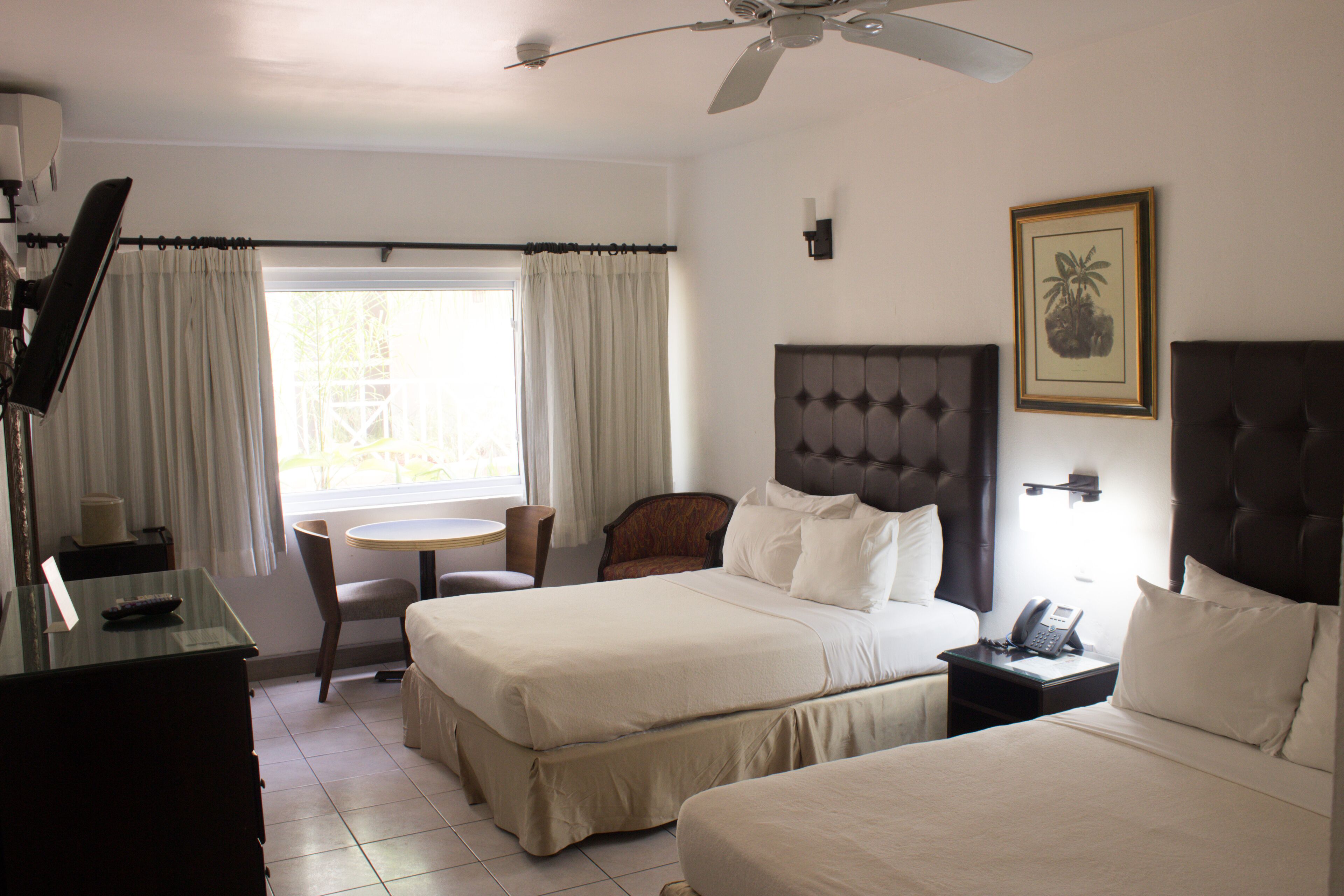 A hotel room with two beds, a desk, a chair, a small table, and a ceiling fan.