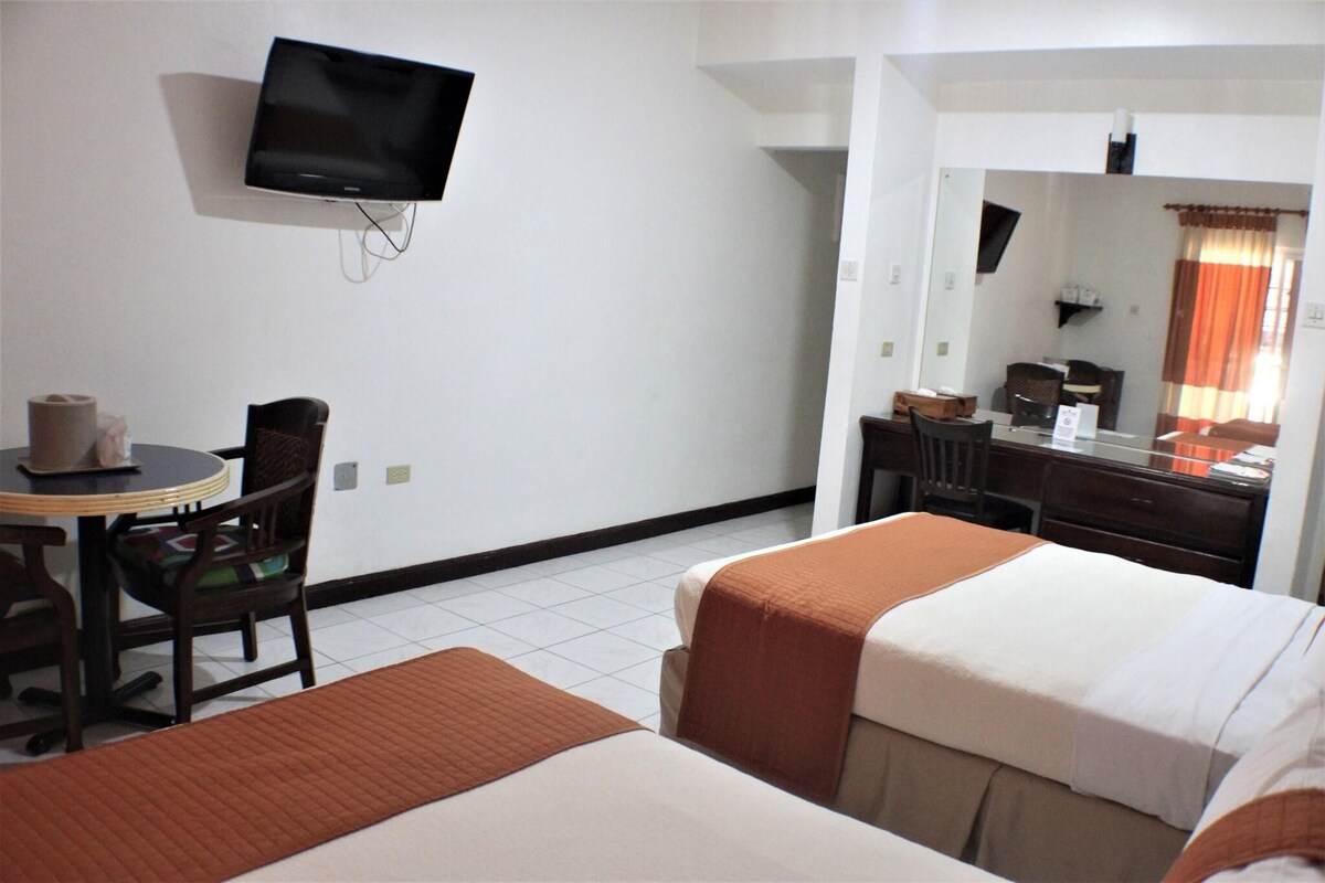 A hotel room with two beds, a TV on the wall, a desk with a chair, and a window with curtains.