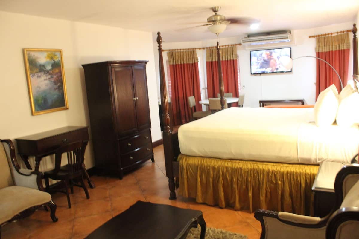 A bedroom with a large bed, a wooden wardrobe, a desk with a chair, and a television on the wall.