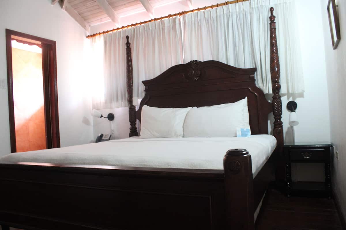 A wooden four-poster bed with white linens, a bedside table with a lamp, and a window with white curtains.