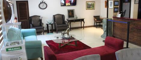 Un hall de hotel con un sofá rojo, dos sillones estampados, una mesa de centro de cristal, un reloj en la pared y una pantalla de televisión.