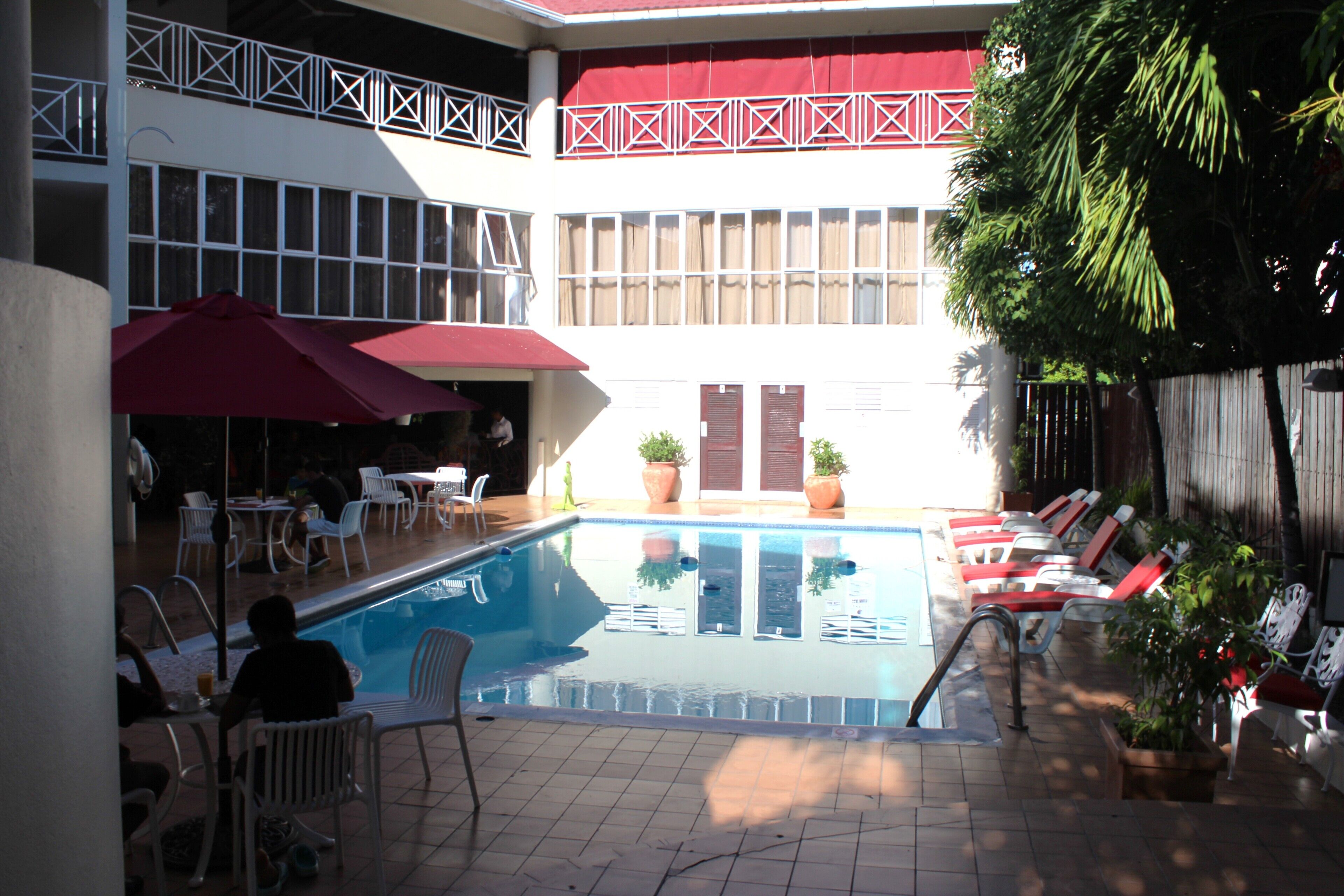 A swimming pool area with lounge chairs and umbrellas, surrounded by potted plants and a building with a patterned balcony.