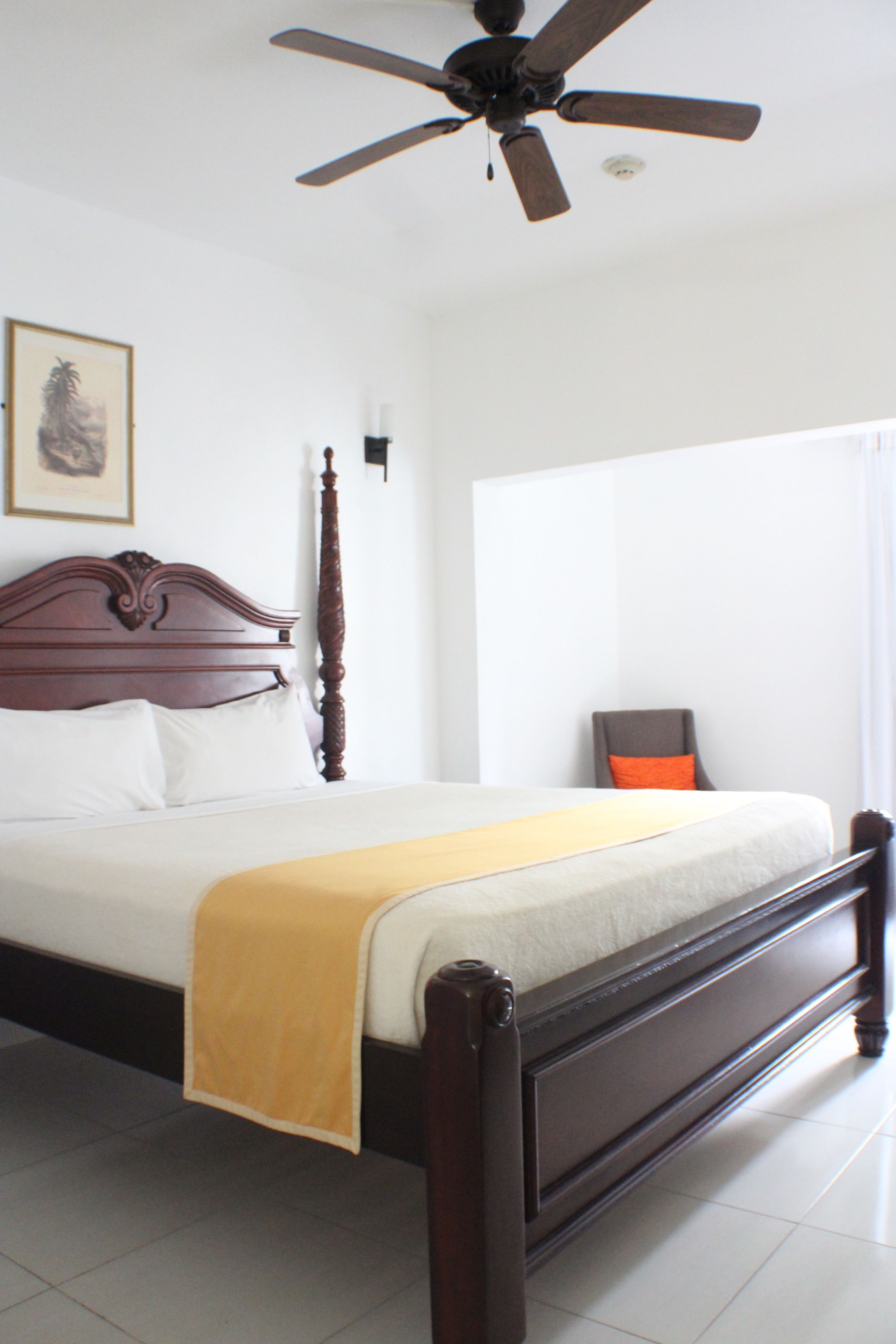 A bedroom with a wooden bed, a ceiling fan, a framed picture on the wall, and a chair.