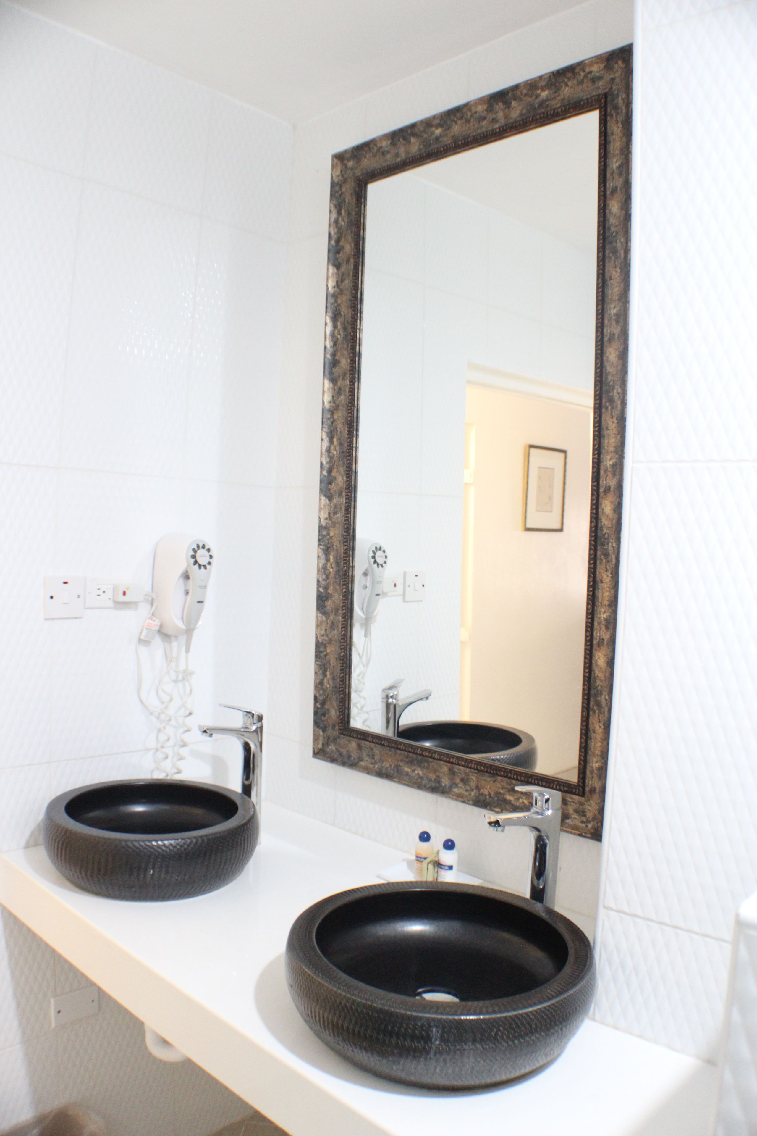 Un baño con dos lavamanos negros, una encimera blanca, un espejo con marco de madera y un secador de pelo fijado a la pared.