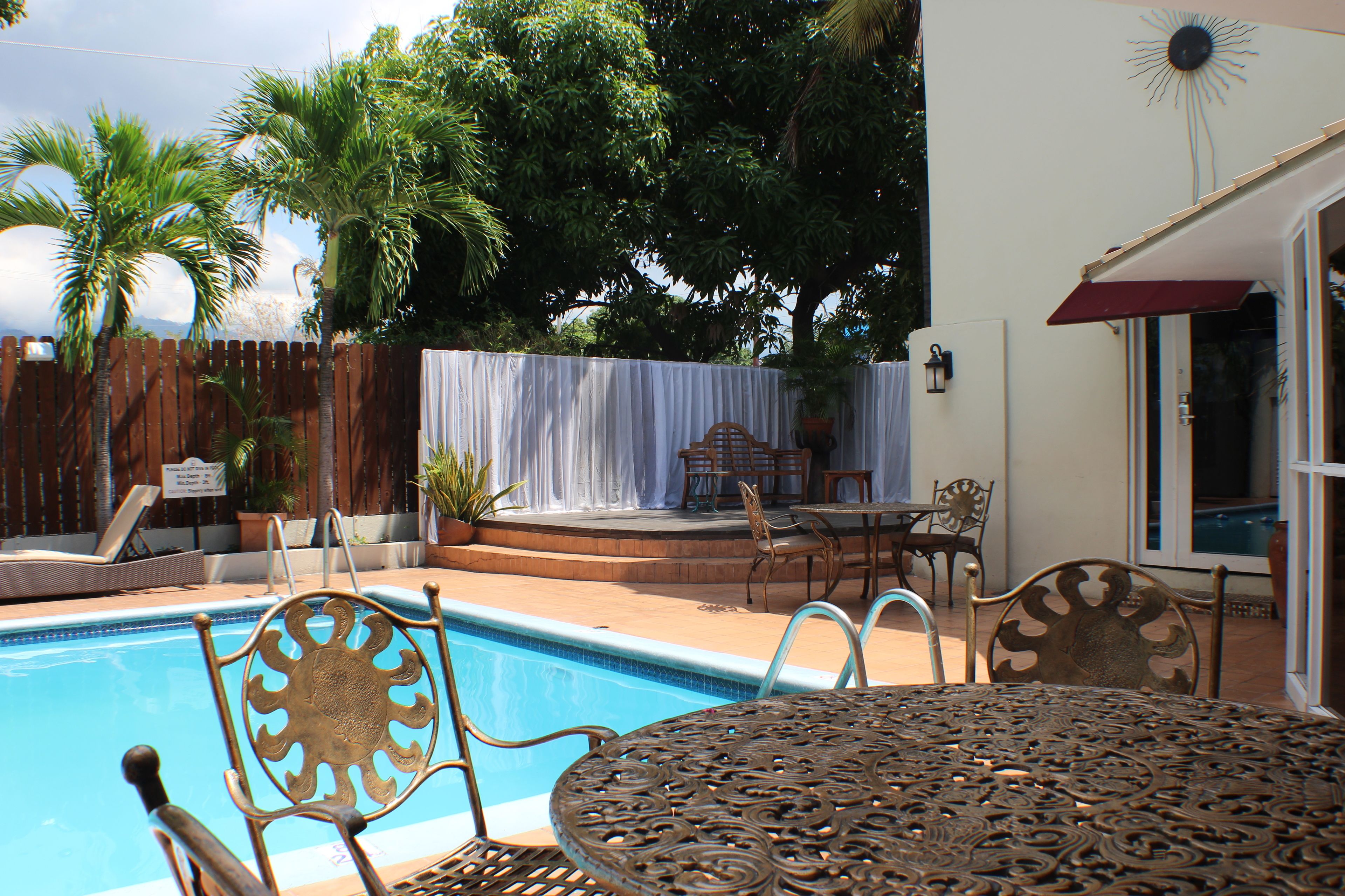 Area piscina con tavolo e sedie in metallo, patio coperto con un palcoscenico e cortile recintato con palme.