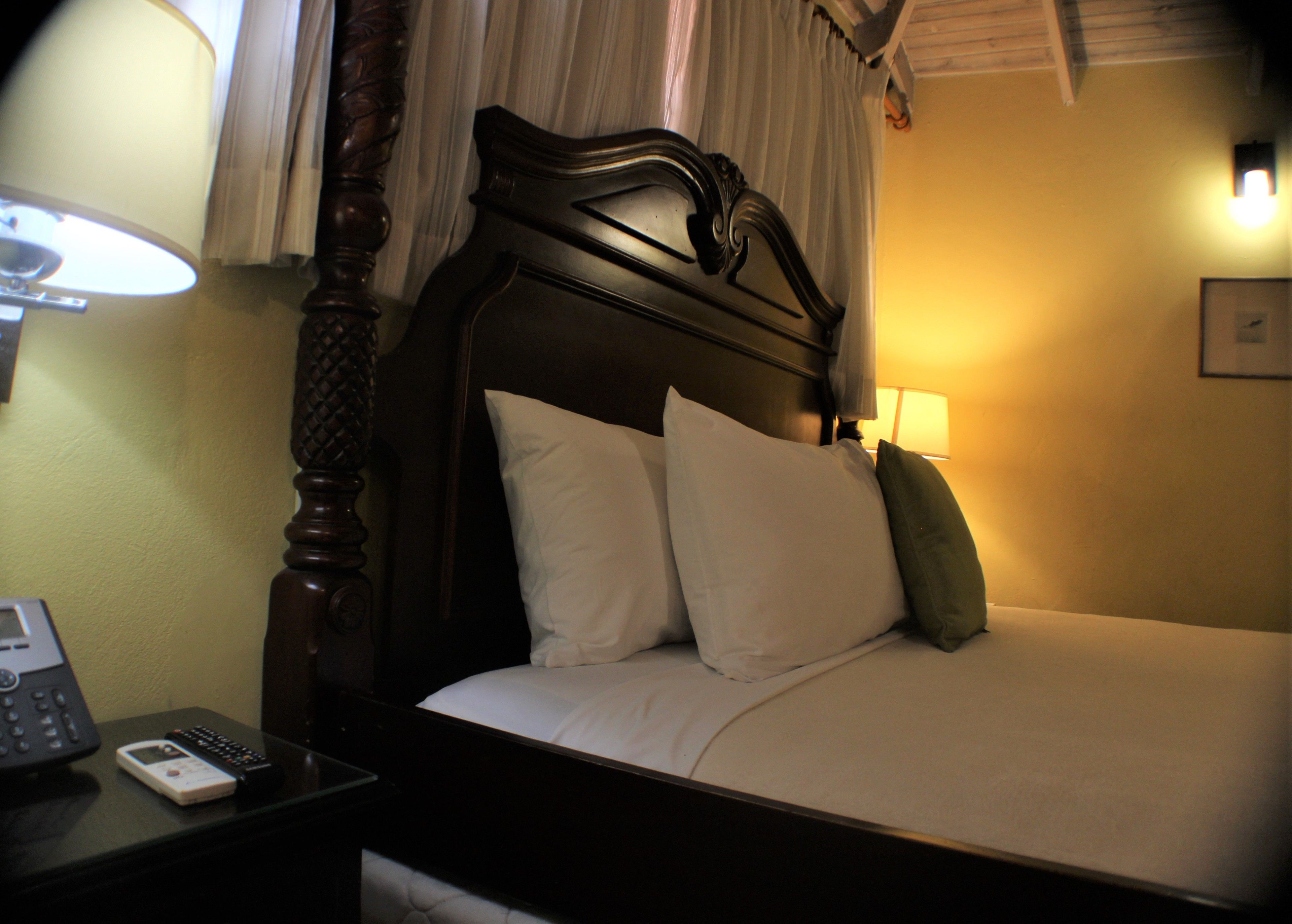 A hotel room with a wooden headboard, a bedside table with a lamp, a phone, and a bed with pillows.
