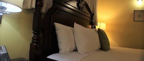 A hotel room with a wooden headboard, a bedside table with a lamp, a phone, and a bed with pillows.