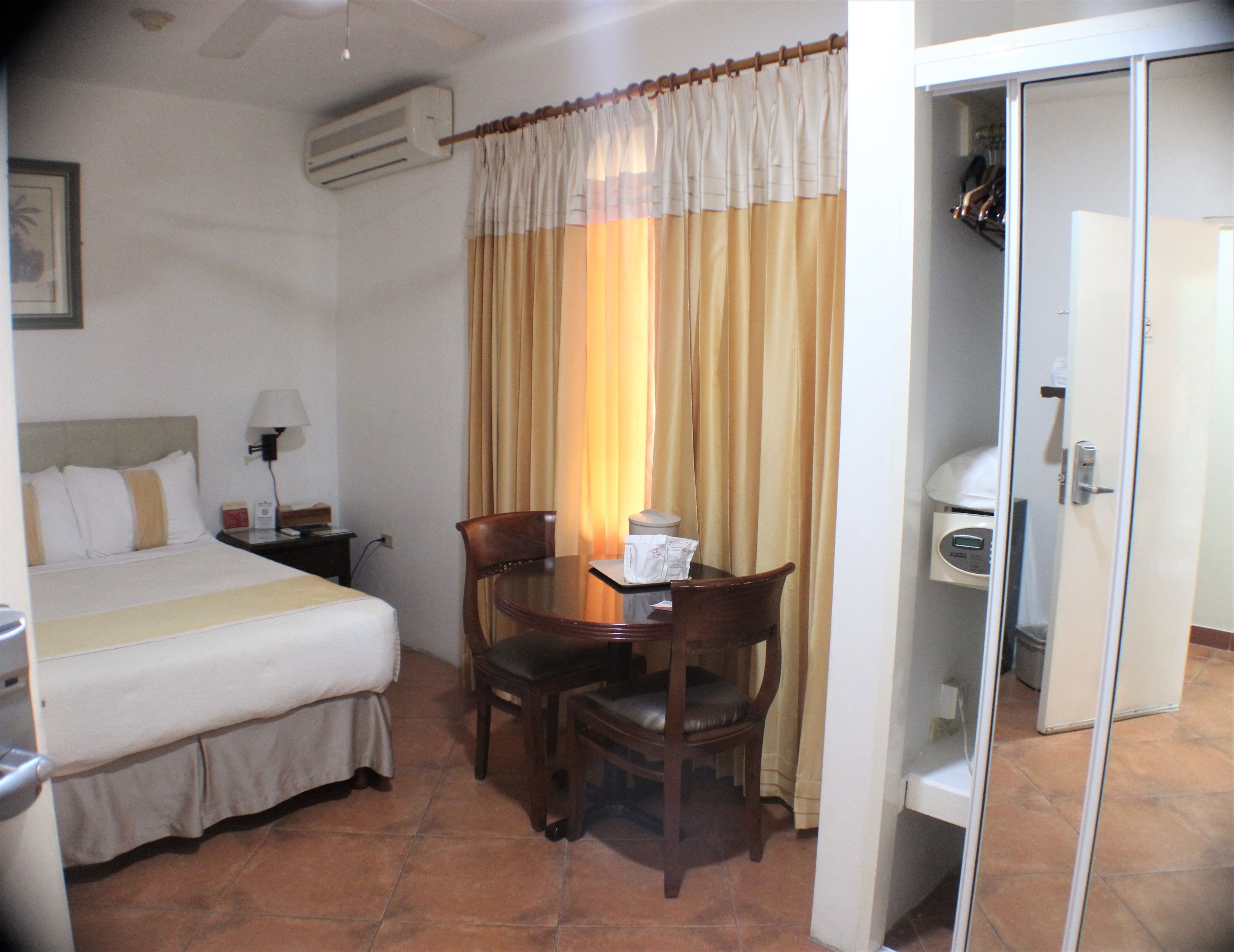 A hotel room with a bed, a desk, a chair, a table, a lamp, a window with curtains, an air conditioning unit, and a door leading to a bathroom.