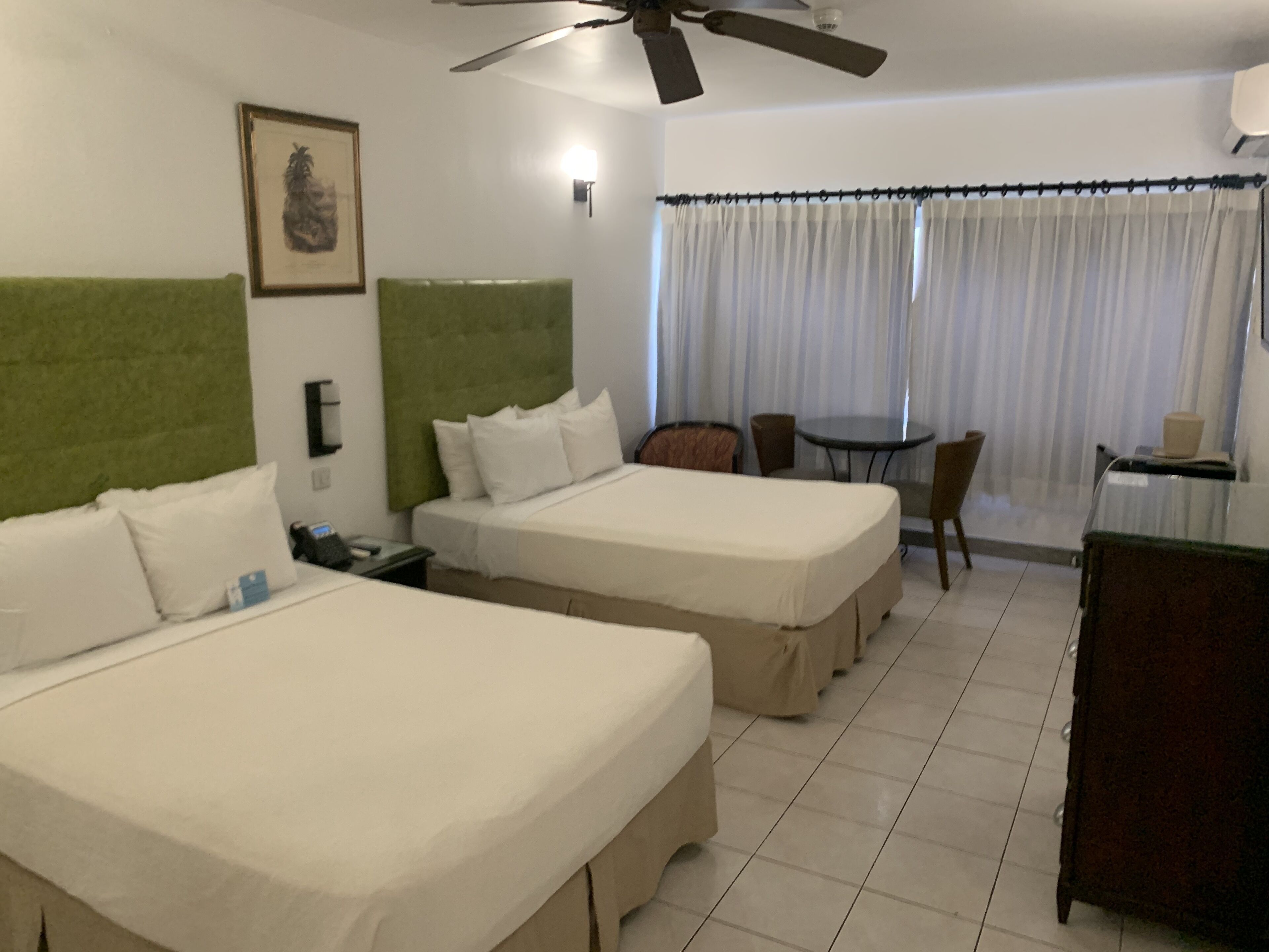 A hotel room with two beds, a green headboard, a desk with a chair, a small table, and a window with curtains.