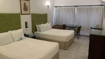 A hotel room with two beds, a green headboard, a desk with a chair, a small table, and a window with curtains.