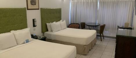 A hotel room with two beds, a green headboard, a desk with a chair, a small table, and a window with curtains.