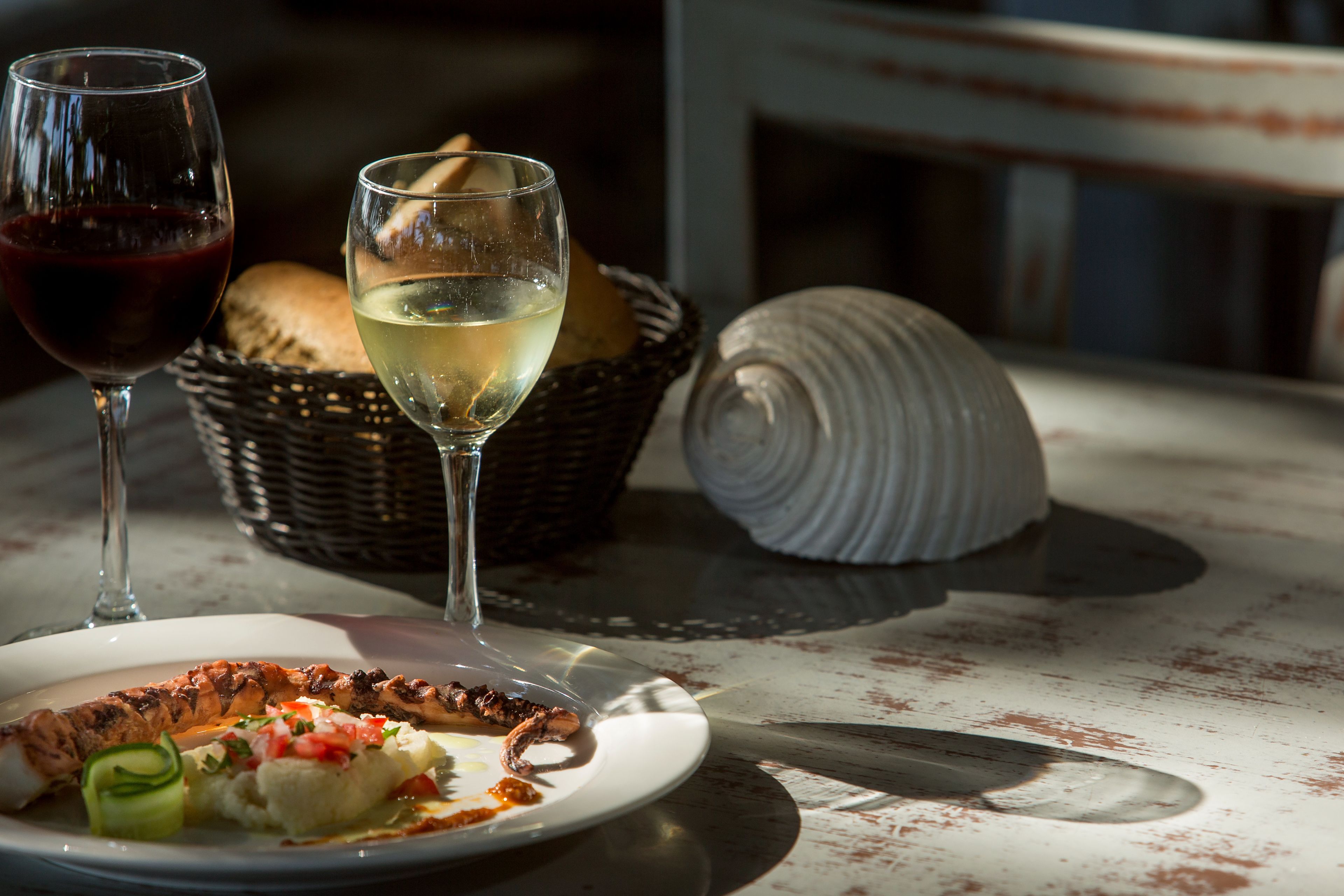 Una mesa con un plato de comida, dos copas de vino, una cesta de pan y una concha marina.