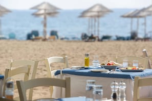A table set for two with a blue tablecloth, white chairs, and a view of the beach and ocean in the background. - Coral Blue Beach Hotel (Polygyros)