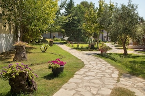 A stone pathway leading through a well-maintained garden with various plants and trees. - Coral Blue Beach Hotel (Polygyros)