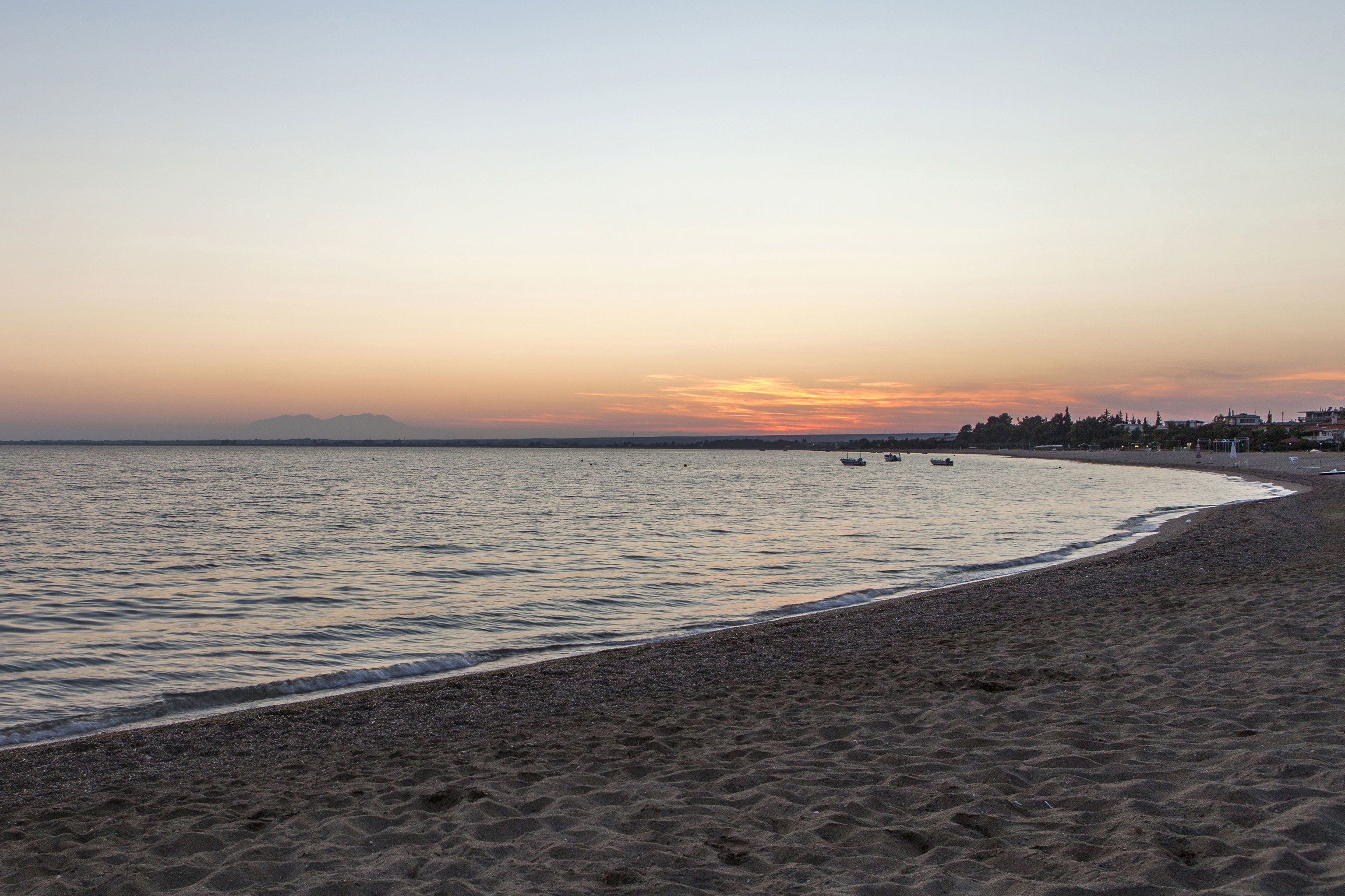 A beach with a calm sea, a sandy shore, and a distant city skyline at sunset.