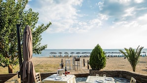 Un área de comedor al aire libre con mesas y sillas, con vistas a una playa con sombrillas y sillones de descanso.
