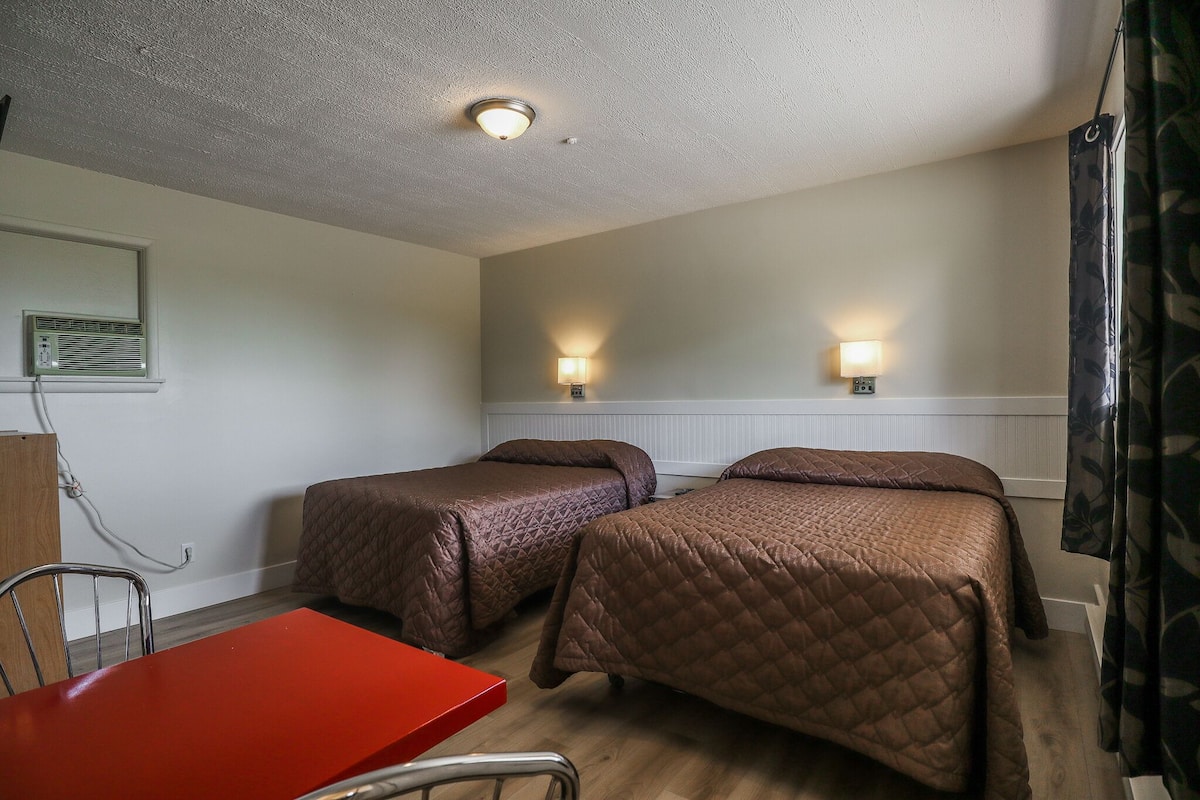 A hotel room with two beds, a red table, a chair, and a ceiling light.