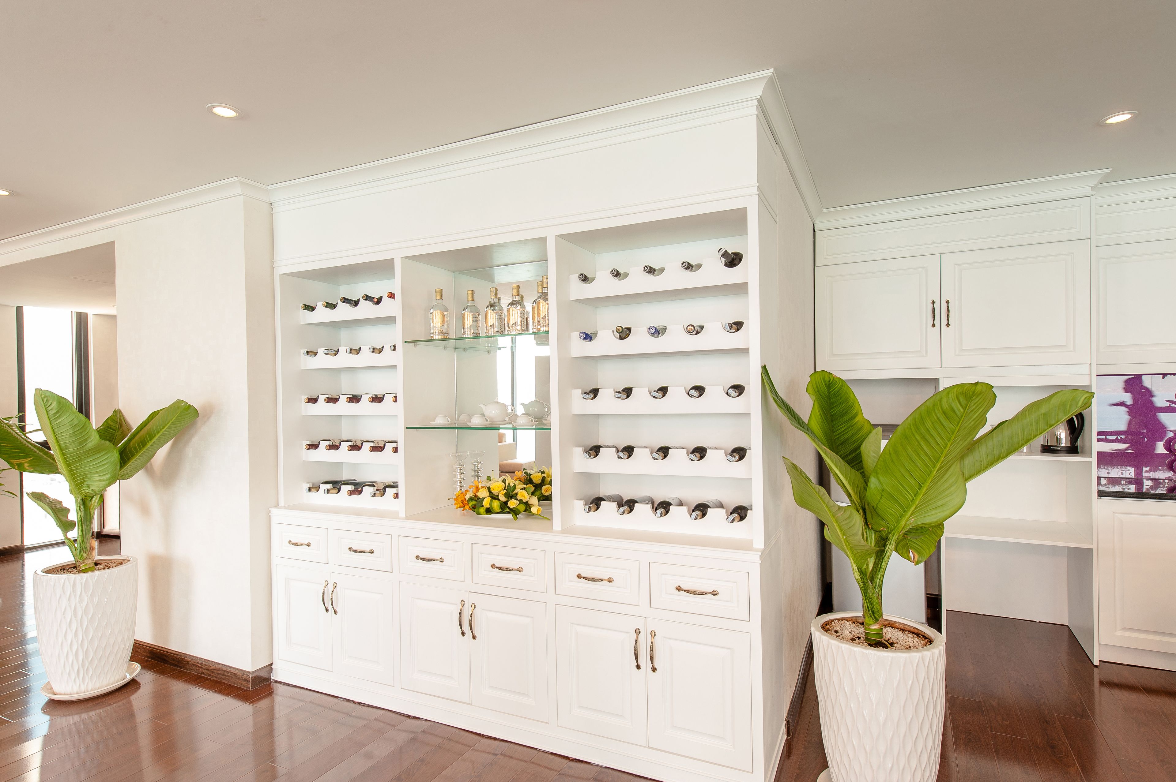 A spacious room with a large white cabinet filled with various items, including bottles and decorative pieces. There are two large potted plants, one on each side of the cabinet.