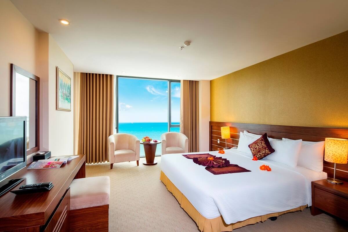 A hotel room with a large bed, a desk with a TV, and a view of the ocean.