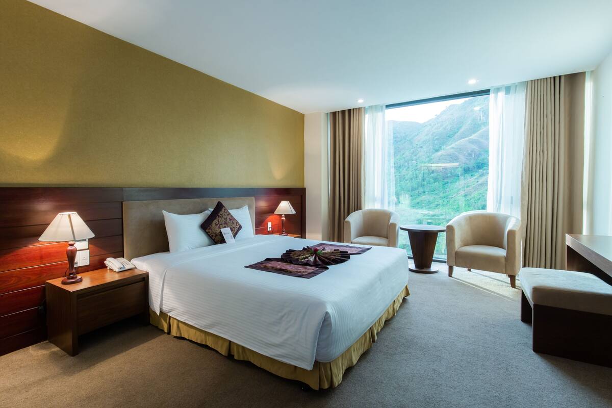 A hotel room with a large bed, two bedside lamps, a nightstand, a desk, two chairs, and a view of a mountainous landscape through large windows.