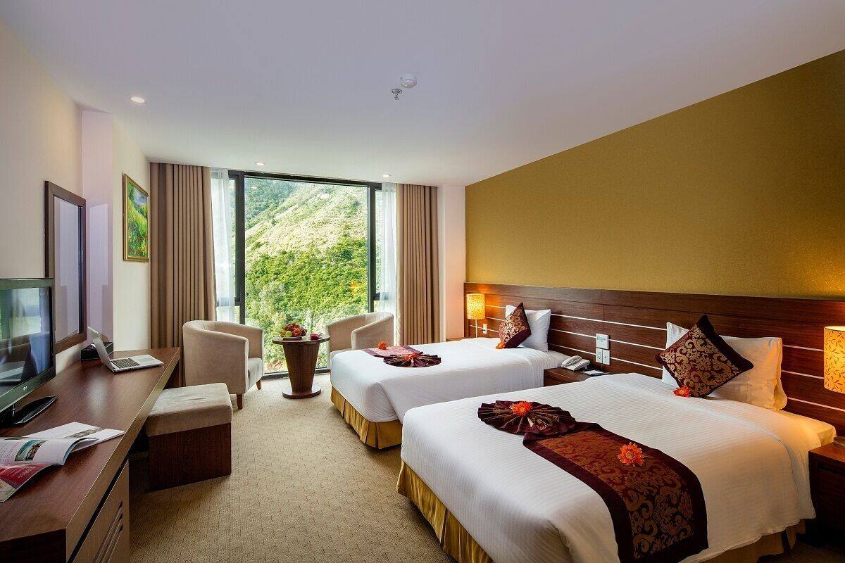 A hotel room with two beds, a desk with a computer, a TV, and a large window offering a view of greenery.