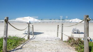 Un sentier en bois mène à une plage où se dressent des parasols blancs, sous un ciel d’un bleu limpide.