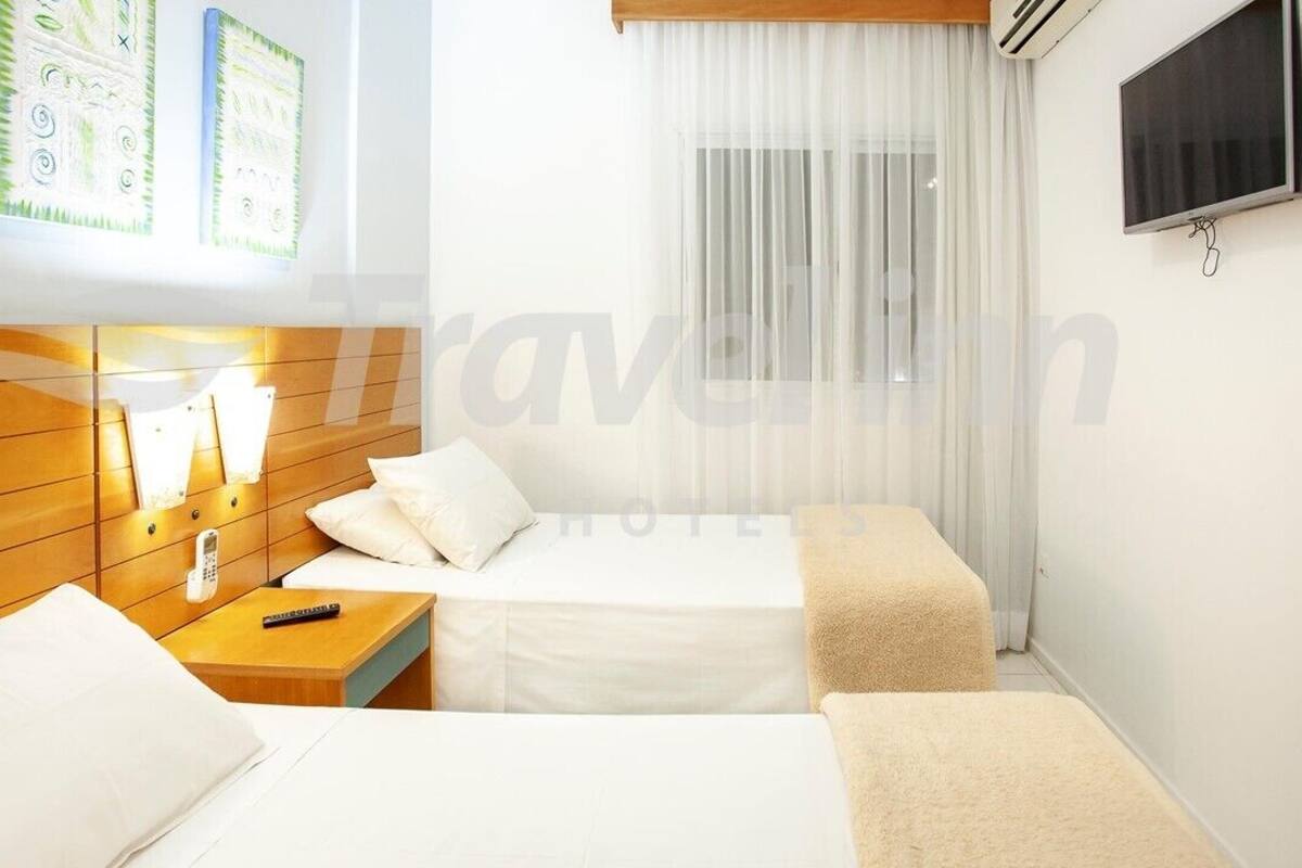A hotel room with two beds, a bedside table with a lamp, a TV mounted on the wall, and artwork on the wall.