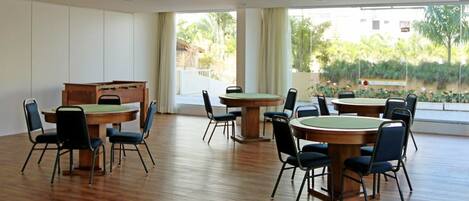 A spacious room with multiple round tables and chairs, large windows with curtains, and a wooden desk.