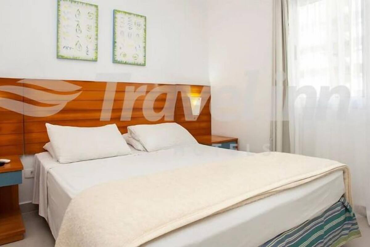 A hotel room with a wooden headboard, a bed with white linens, two bedside tables, and a window with curtains.